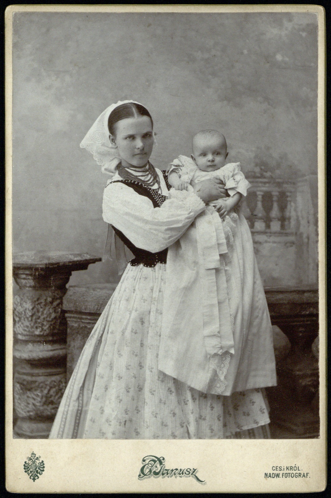 Młoda kobieta w stroju rzeszowskim — haftowana koszula, aksamitny gorset, korale, czepiec — z niemowlęciem na rękach
